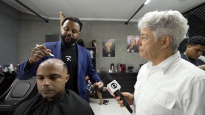 Caco Barcellos segura microfone da TV Globo enquanto entrevista barbeiro que aplica prótese capilar em cliente careca no Profissão Repórter