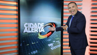 Reinaldo Gottino sorri e gesticula com as duas mãos em direção ao telão com o logotipo do Cidade Alerta no estúdio da Record