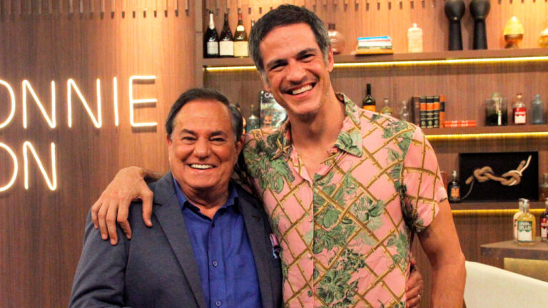 Ronnie Von e Mateus Solano sorrindo e posando juntos em um estúdio com decoração sofisticada e prateleiras com livros e bebidas ao fundo