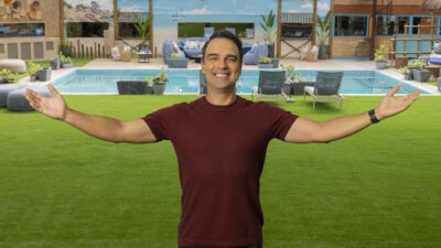 Tadeu Schmidt de braços abertos no jardim da casa do BBB 25 com piscina e decoração ao fundo