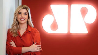 Thalita Vieira, nova diretora de Marketing da Jovem Pan, posa encostada em uma parede branca, usando camisa vermelha e calça preta. Ao fundo, o logotipo luminoso da Jovem Pan em uma parede vermelha