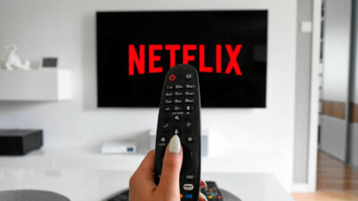 Mão de assinante segurando controle remoto apontado para uma TV que exibe o logotipo da Netflix em uma sala moderna