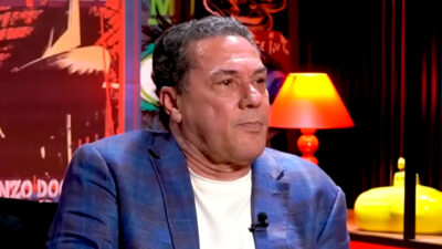 Vanderlei Luxemburgo durante entrevista, vestindo blazer azul e camiseta branca, com cenário colorido e luminária acesa ao fundo