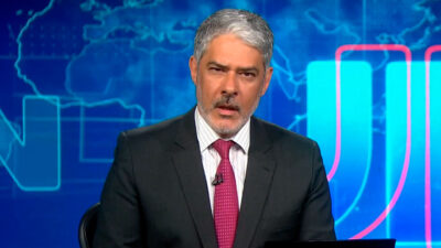 William Bonner apresenta o Jornal Nacional no estúdio, vestindo terno escuro e gravata rosa, com fundo azul exibindo mapa-múndi estilizado