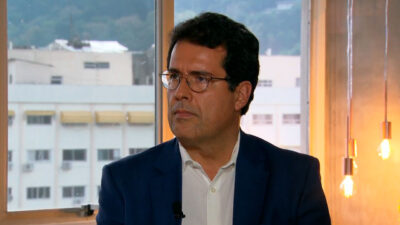André Trigueiro, de terno azul e óculos, aparece sério durante uma entrevista, com janelas ao fundo mostrando prédios e montanhas