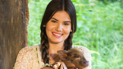 Camila Queiroz caracterizada como Mafalda em Êta Mundo Bom!, sorrindo e segurando um leitão com um laço no pescoço, vestindo blusa rendada e com cabelos trançados, diante de um cenário rural verdejante