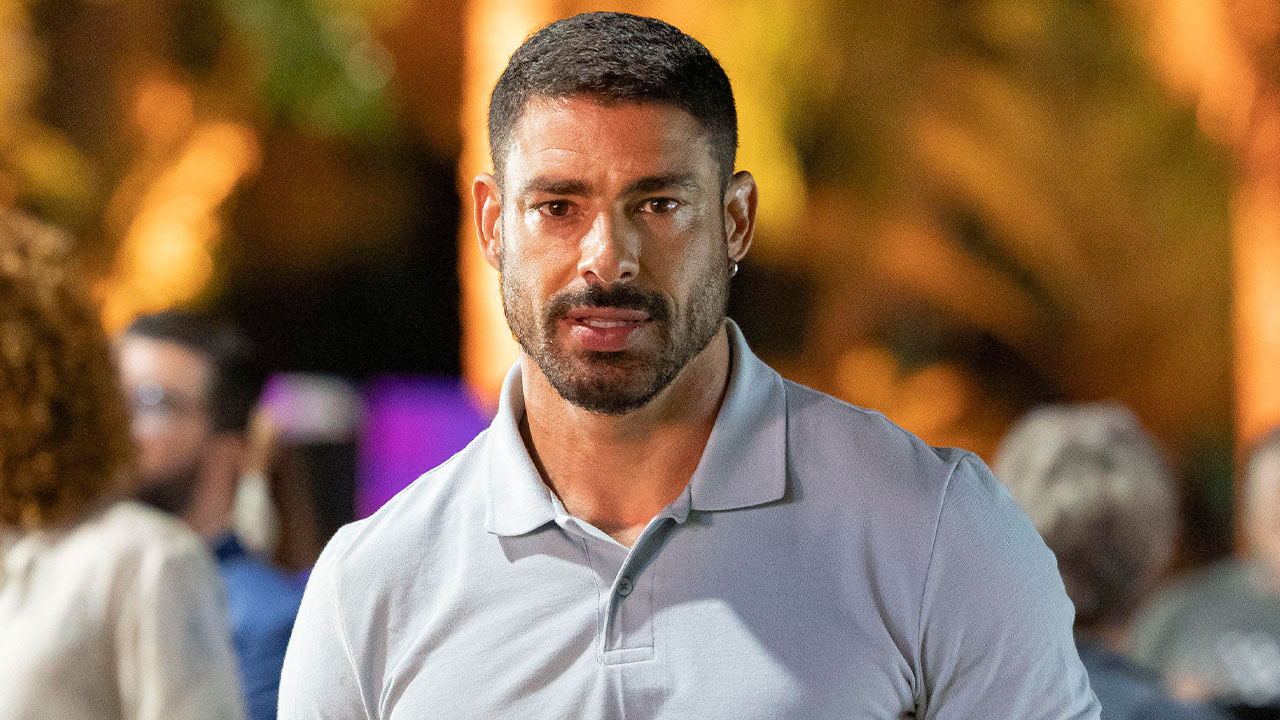 Cauã Reymond interpretando César Ribeiro no remake de Vale Tudo, com expressão séria em uma cena noturna ao ar livre, com pessoas ao fundo