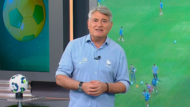 Cléber Machado no estúdio da Record durante transmissão de futebol com tela de jogo ao fundo e bola sobre bancada