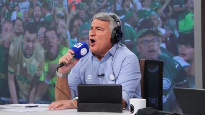 Cléber Machado narrando um jogo na Record, segurando um microfone e usando fones de ouvido, com uma torcida vibrante ao fundo