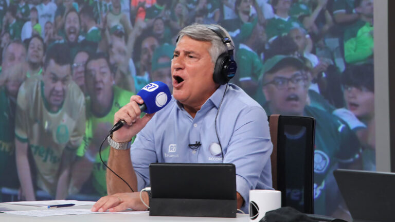 Cléber Machado narrando um jogo na Record, segurando um microfone e usando fones de ouvido, com uma torcida vibrante ao fundo