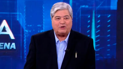 Datena apresentando o programa Tá na Hora, vestindo terno escuro e camisa azul, com fundo azul e letreiro do programa ao fundo