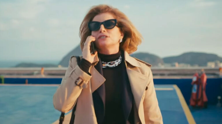 Débora Bloch caracterizada como Odete Roitman no remake de Vale Tudo, usando óculos escuros e casaco bege enquanto fala ao celular em um heliponto, com o Pão de Açúcar ao fundo