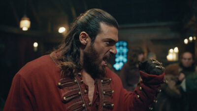 Can Yaman caracterizado como seu personagem na série El Turco, vestindo traje vermelho e expressando intensidade em uma cena