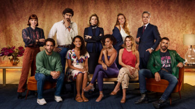 Elenco do remake de Vale Tudo posando para foto oficial, com atores distribuídos entre posições sentadas e em pé, em um cenário sofisticado com tons dourados e avermelhados
