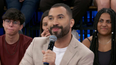 Gil do Vigor segura um microfone enquanto fala no programa Altas Horas, vestindo um blazer bege e camisa branca, com plateia ao fundo
