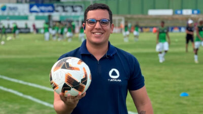 Ícaro Novaes, repórter da TV Atalaia, afiliada da Record, segura uma bola de futebol em campo durante a cobertura de um jogo