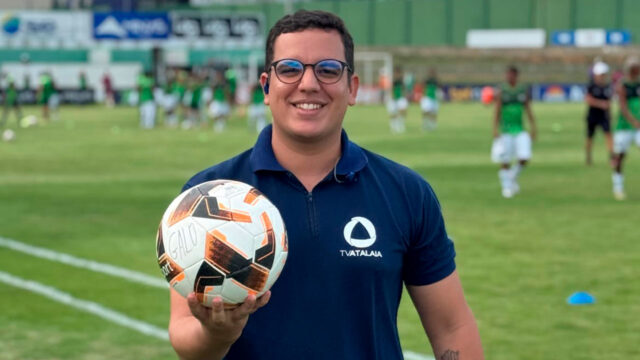 Ícaro Novaes, repórter da TV Atalaia, afiliada da Record, segura uma bola de futebol em campo durante a cobertura de um jogo