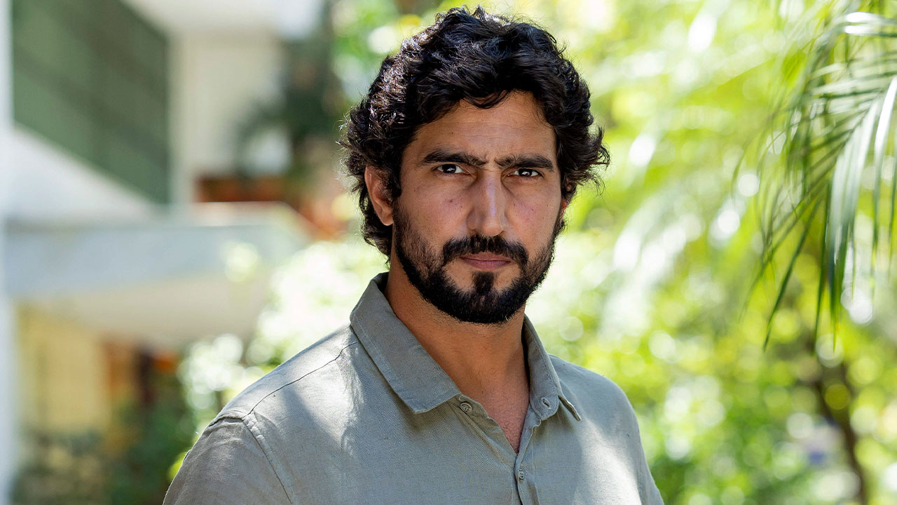 Renato Góes interpretando Ivan Meireles no remake de Vale Tudo, com expressão séria em um cenário ao ar livre com vegetação ao fundo