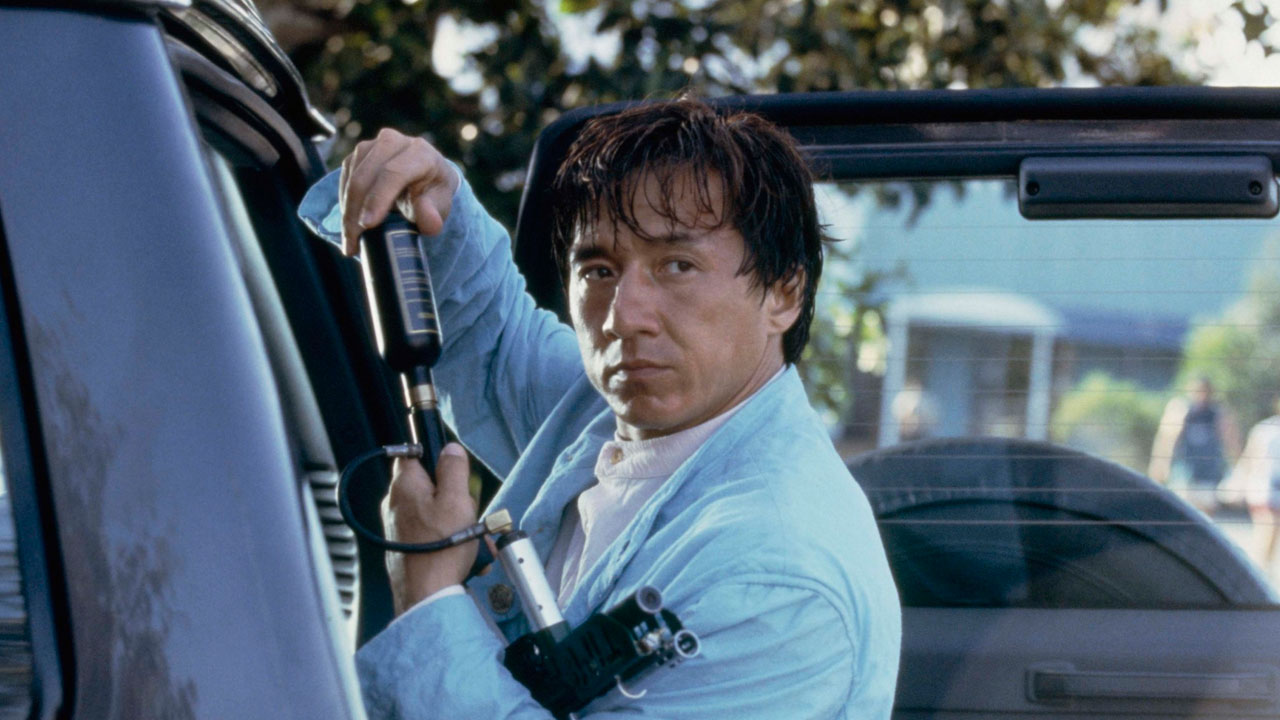 Jackie Chan em cena de Primeiro Impacto segurando uma arma improvisada enquanto usa um paletó azul claro ao lado de um carro