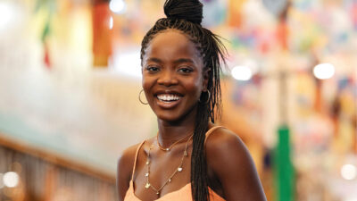 Clara Moneke caracterizada como Leona, personagem principal da novela Dona de Mim, sorrindo em um ambiente iluminado e colorido