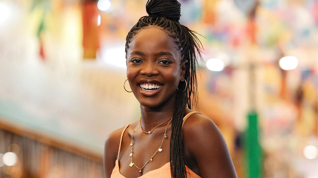 Clara Moneke caracterizada como Leona, personagem principal da novela Dona de Mim, sorrindo em um ambiente iluminado e colorido