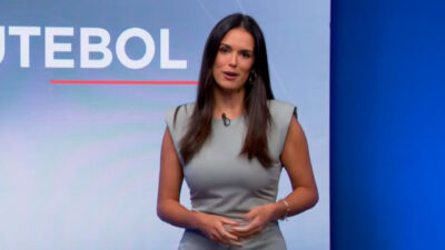 Lívia Nepomuceno apresentando o Jornal da Band, vestindo um vestido cinza justo, com fundo azul e letreiro parcial com a palavra "Futebol" ao fundo