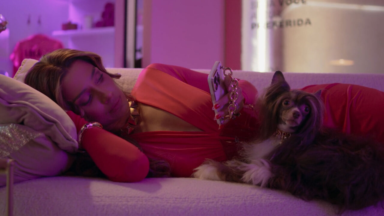Camila Pitanga, no papel de Lola, está deitada em um sofá segurando o celular, vestindo roupa vermelha e acessórios dourados, com o cachorro Lolindo ao seu lado, em cena da novela Beleza Fatal
