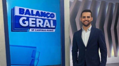 Lucas Carvalho sorri enquanto posa no estúdio do Balanço Geral SP Campinas-Manhã, vestindo terno azul e camisa clara, ao lado do telão com o logotipo do programa
