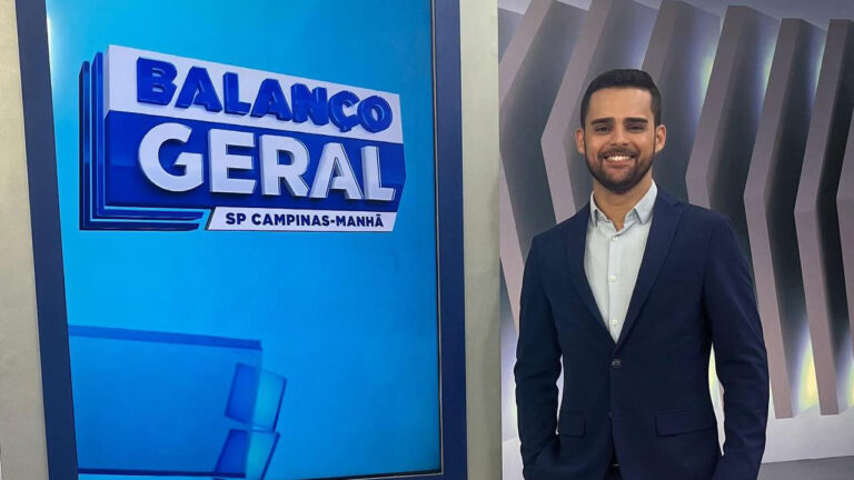 Lucas Carvalho sorri enquanto posa no estúdio do Balanço Geral SP Campinas-Manhã, vestindo terno azul e camisa clara, ao lado do telão com o logotipo do programa