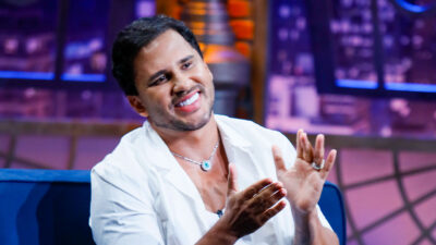 Lucas Guimarães durante entrevista no The Noite, sorrindo e aplaudindo enquanto veste camisa branca e colar com pedra azul, com cenário iluminado ao fundo