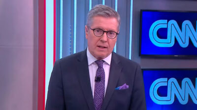 Márcio Gomes apresentando o CNN Prime Time da CNN Brasil, usando terno escuro e óculos, com o logotipo do canal ao fundo