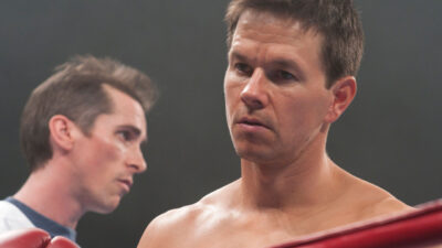 Mark Wahlberg, de luvas de boxe, olha sério dentro do ringue, enquanto Christian Bale aparece desfocado ao fundo, em cena do filme O Vencedor