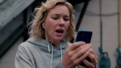 Naomi Watts como Amy aparece aflita enquanto olha para o celular, usando moletom cinza e fones de ouvido, em cena do filme A Hora do Desespero