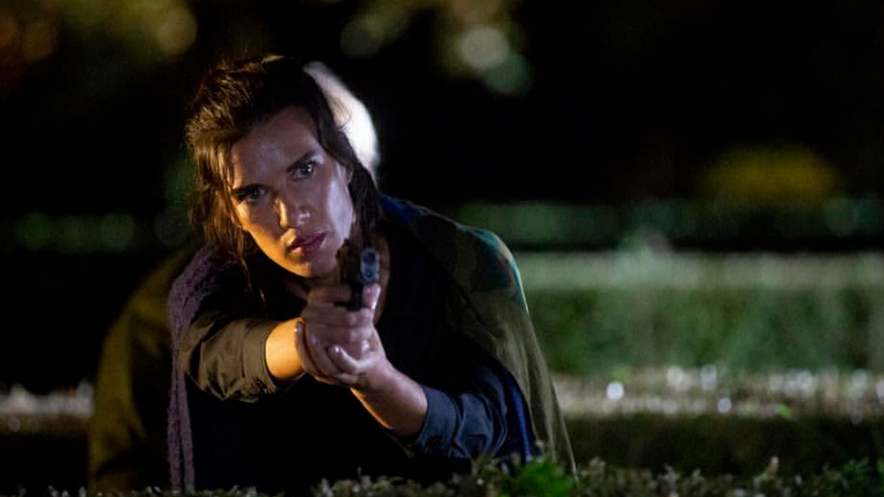 Natalie Burn como Emma aponta uma arma com expressão determinada enquanto se esconde atrás de uma cerca viva durante a noite no filme Sobreviva à Noite (2019)