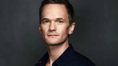 Neil Patrick Harris posa para foto com expressão séria, vestindo camisa preta, diante de um fundo escuro