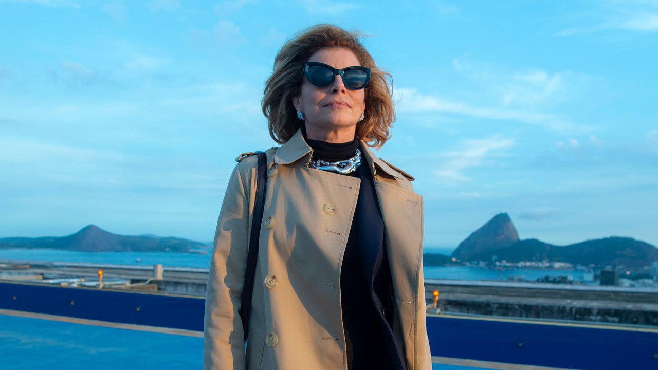 Debora Bloch interpretando Odete Roitman no remake de Vale Tudo, usando óculos escuros e sobretudo bege, com o Pão de Açúcar ao fundo