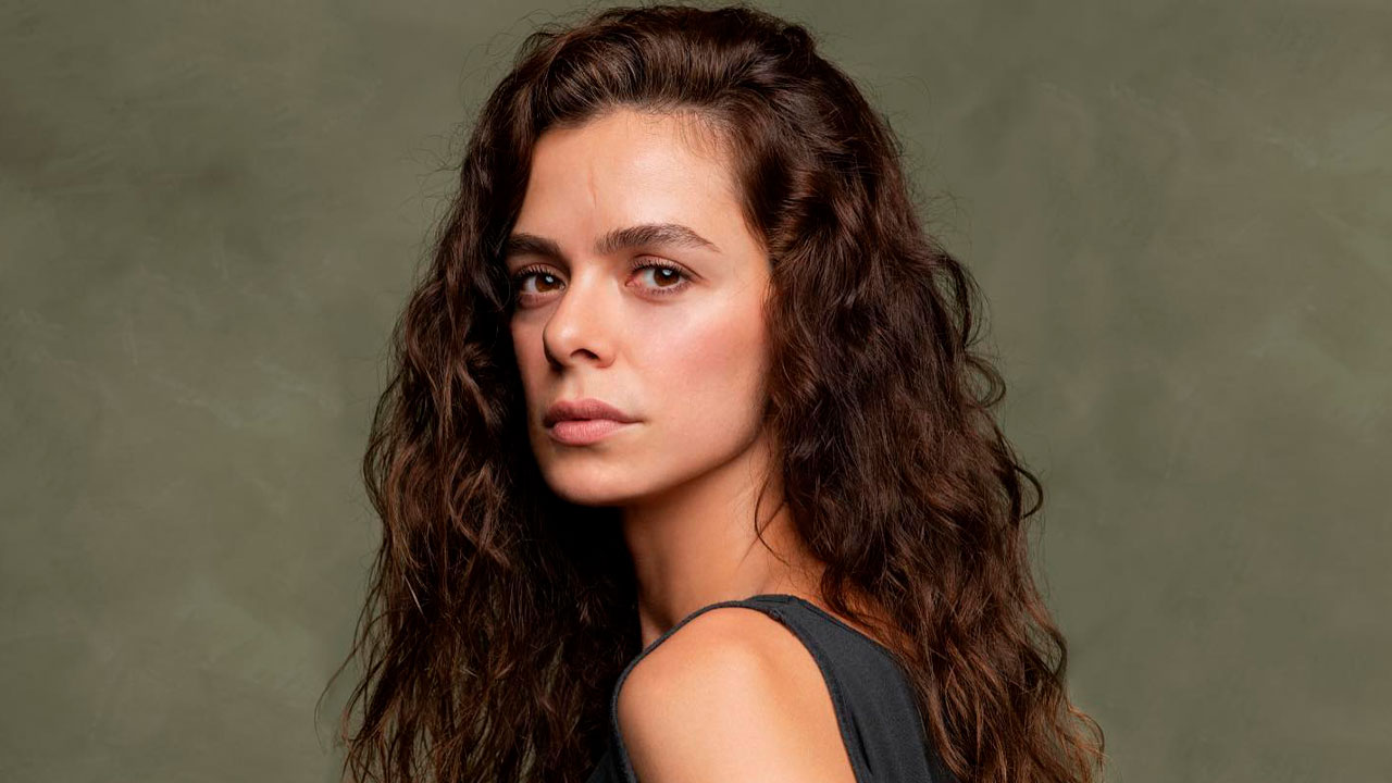 Özge Özpirinçci em ensaio fotográfico, com cabelo cacheado solto, usando blusa preta e olhando séria para a câmera, sobre fundo verde neutro