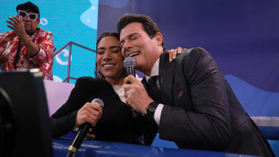 Patricia Abravanel e Celso Portiolli cantam juntos no palco do Teleton 2024 segurando microfones e abraçados, enquanto ao fundo Liminha anima o público com óculos escuros e roupa vermelha