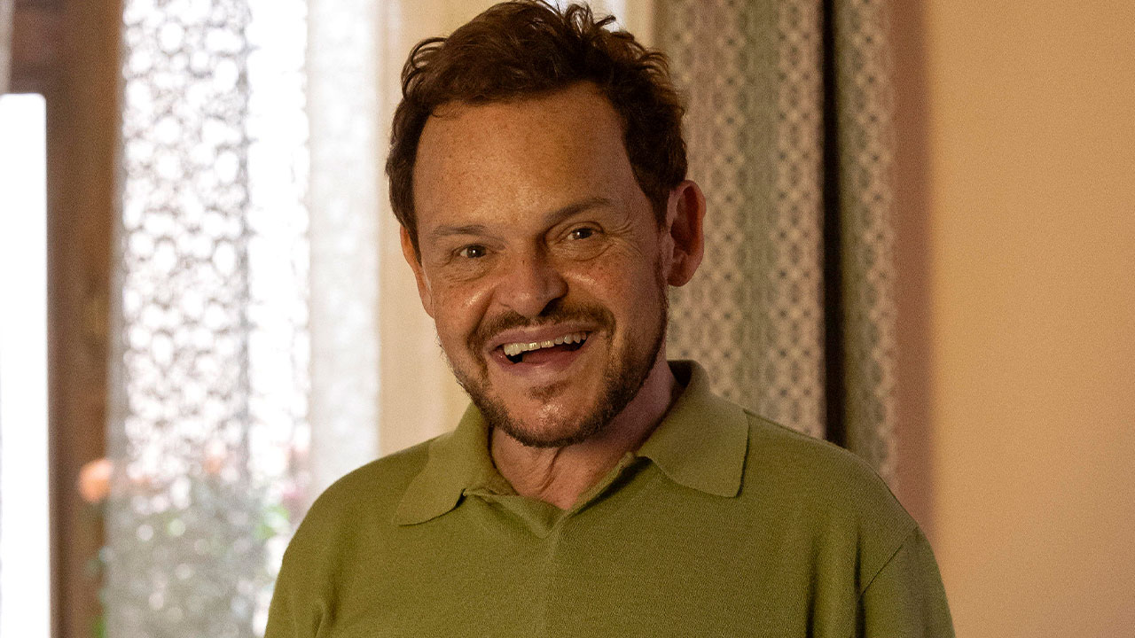 Matheus Nachtergaele interpretando Poliana no remake de Vale Tudo, sorrindo em um ambiente interno com cortinas decorativas ao fundo
