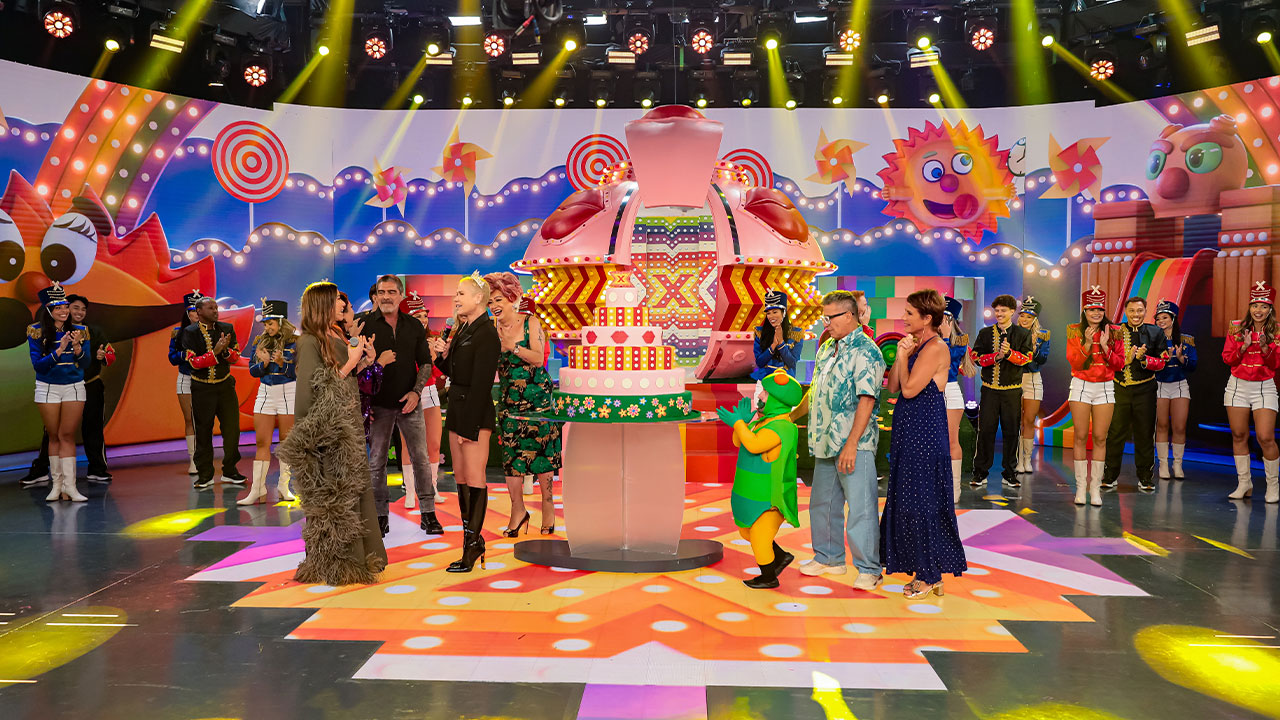 Xuxa Meneghel comemora no palco do Programa Silvio Santos com bolo temático e convidados, cenário colorido e personagens infantis ao fundo