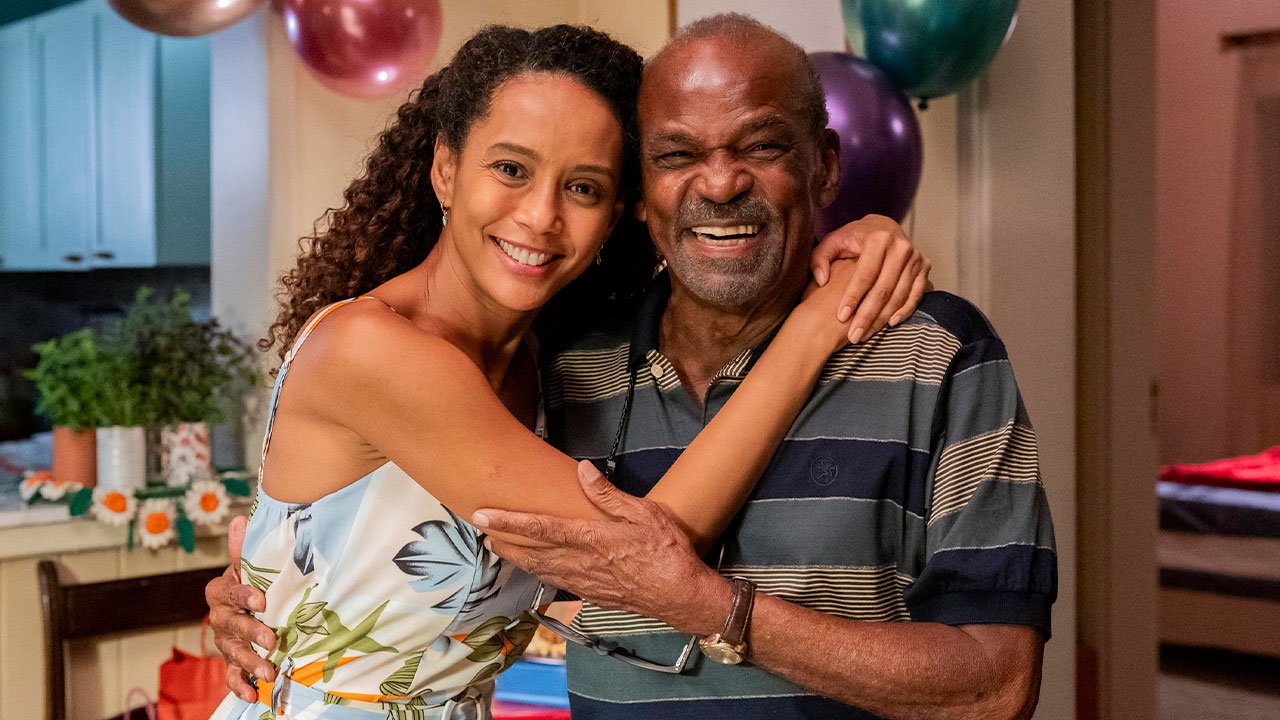 Taís Araújo como Raquel abraçando Antonio Pitanga como Salvador em cena do remake de Vale Tudo, sorrindo em um ambiente decorado com balões