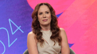 Regina Volpato sentada em um estúdio de TV, vestindo um vestido claro, com cabelo solto e ondulado, diante de um fundo colorido com tons de rosa e roxo