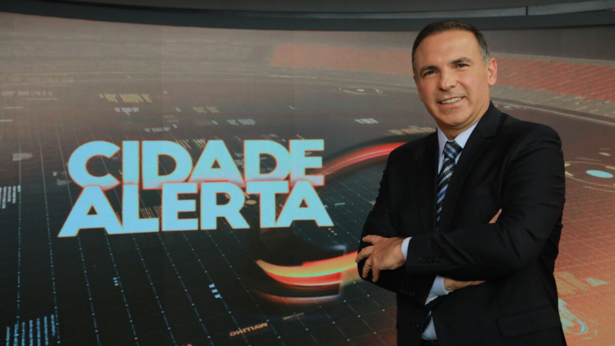 Reinaldo Gottino sorrindo no estúdio do Cidade Alerta, de braços cruzados, com o logotipo do programa ao fundo