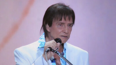 Roberto Carlos segura um microfone enquanto canta no palco, vestindo um terno branco com camisa azul estampada, diante de um fundo claro