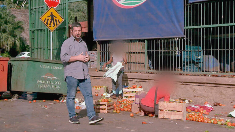 Rafael Batalha, repórter do SBT, em local da Ceagesp com caixas de tomates espalhadas no chão e pessoas recolhendo os alimentos na série especial Depósito do Crime