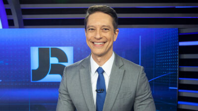 Sérgio Aguiar sorrindo no estúdio do Jornal da Record, vestindo terno cinza e gravata azul, com o logotipo do telejornal ao fundo