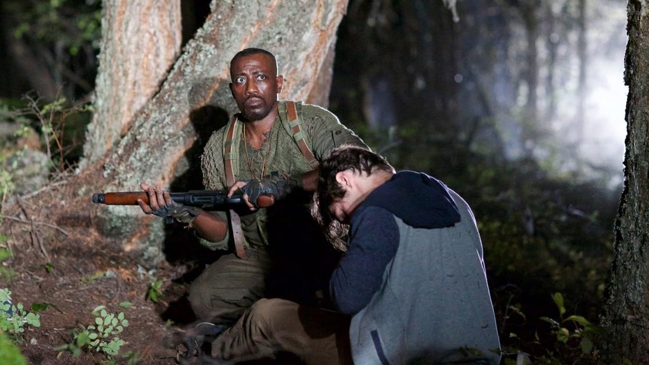 Wesley Snipes segura uma espingarda enquanto se protege atrás de uma árvore com RJ Mitte, que está abaixado, no filme Invasão Alienígena (2017)