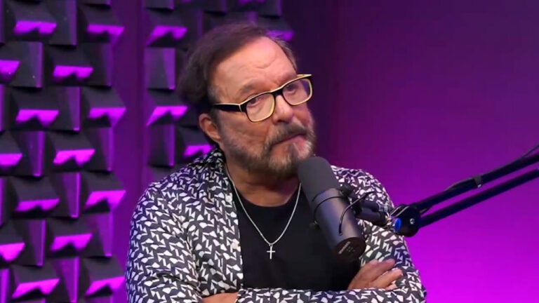 Wolf Maya durante entrevista no estúdio do podcast Papagaio Falante, usando óculos de armação amarela, camisa estampada em preto e branco e colar com crucifixo, com iluminação roxa ao fundo
