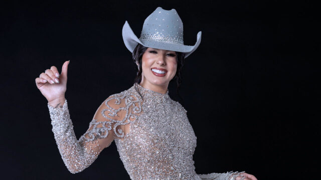 Cantora Ana Castela sorrindo com vestido brilhante e chapéu prateado em ensaio com fundo preto