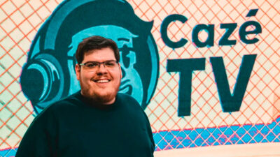 Casimiro Miguel sorri em frente a painel com logo e nome do canal CazéTV ao fundo